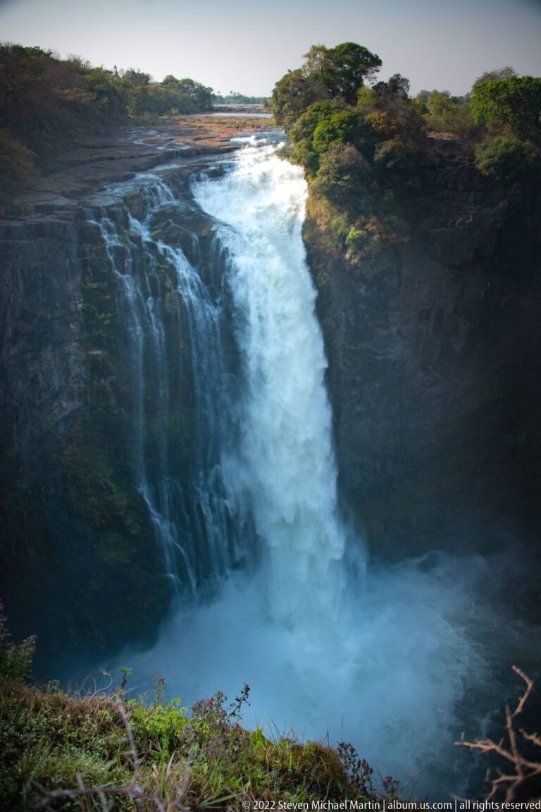 Concentrated waterfall plunging from a flat rocky lip into a mist-filled basin bordered by dark basalt and vegetation.