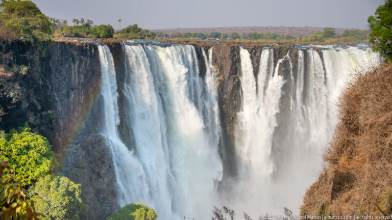 Broad frontal curtain of Victoria Falls descending in parallel streams over a dark basalt rim into dense spray.