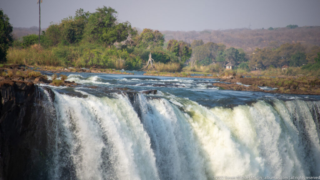 Upper Zambezi River rushing over the brink of Victoria Falls, with rapids and wooded riverbank visible beyond the crest.
