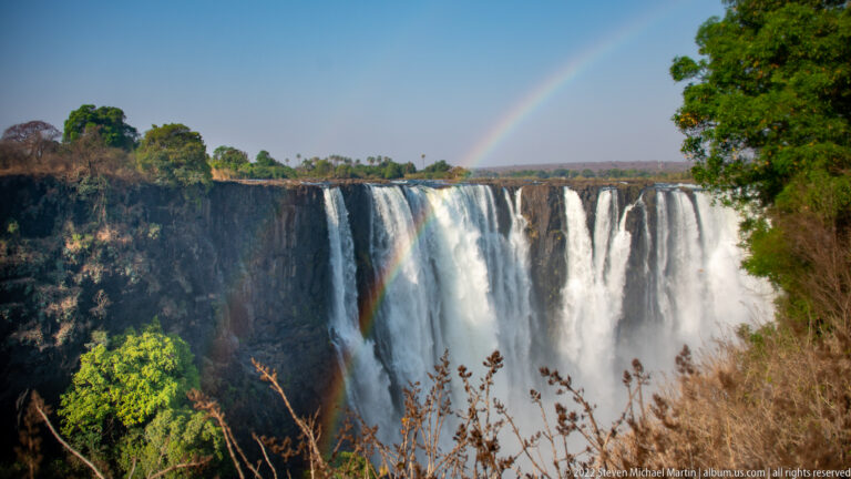 Bright rainbow descending beside segmented white curtains of Victoria Falls, with basalt cliff and green rim vegetation.
