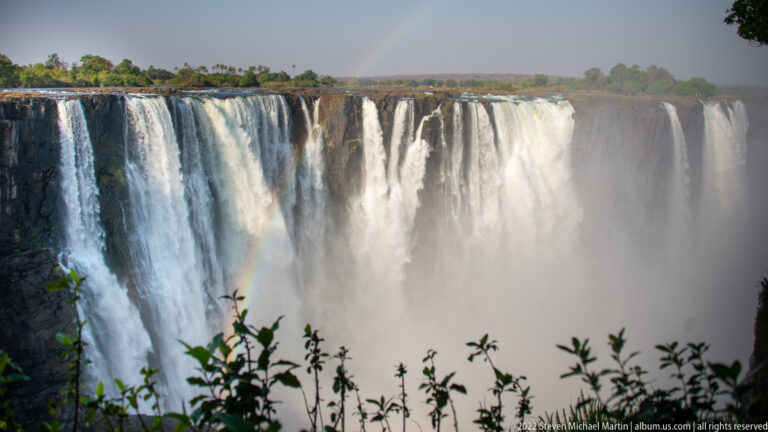 Frontal view of tall parallel waterfalls at Victoria Falls, with heavy mist, a faint rainbow, and leaves in the foreground.