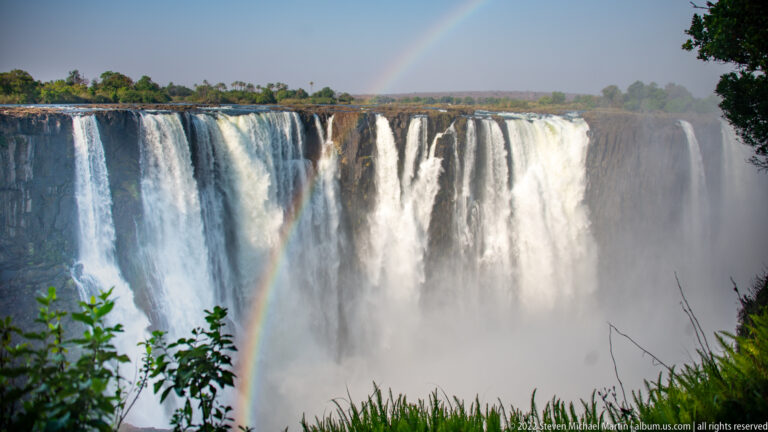 Broad section of Victoria Falls with a narrow rainbow rising from mist and leafy shrubs framing the foreground.