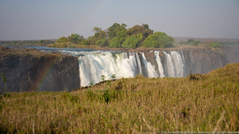 Wide view of Victoria Falls beyond tawny dry-season grass, with several waterfall curtains and a green tree-covered island.