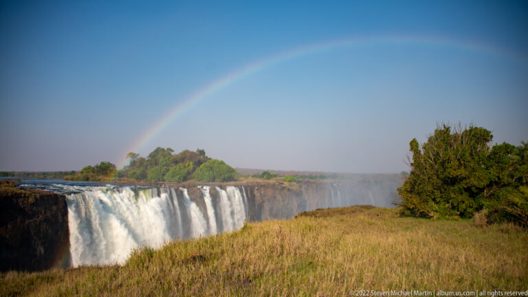 Full rainbow arching above Victoria Falls, with white cascades, a green island, and golden dry-season grass in the foreground.