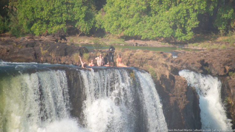 Several people standing and sitting on exposed rock beside flowing water at the lip of Victoria Falls.