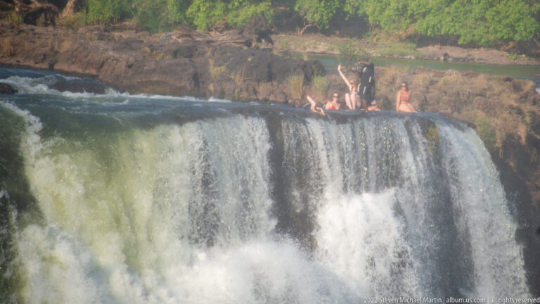 People gathered on dark rock just above the waterfall edge, with white water dropping in the foreground and a side cascade at right.