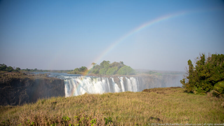 Distant section of Victoria Falls beneath a pale rainbow, seen across open dry grassland with scrub at right.
