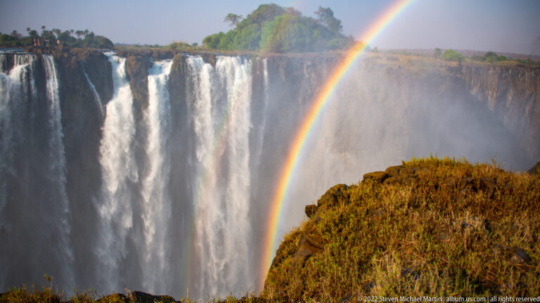 Close rainbow rising through waterfall mist beside a tall white curtain of Victoria Falls and a sunlit grassy rock outcrop.