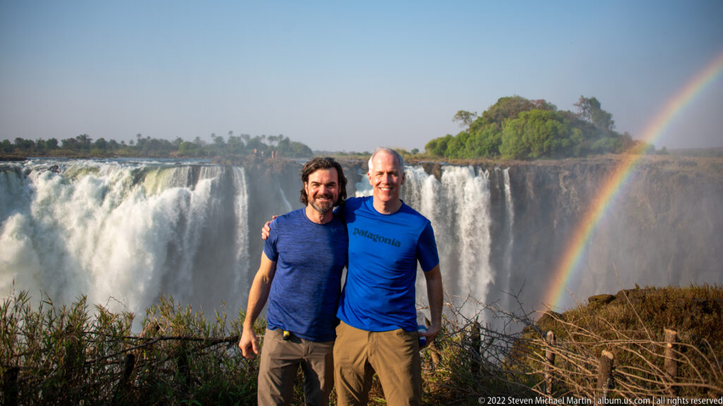 Two men posing at a Victoria Falls viewpoint, with a broad waterfall curtain and bright rainbow behind them.