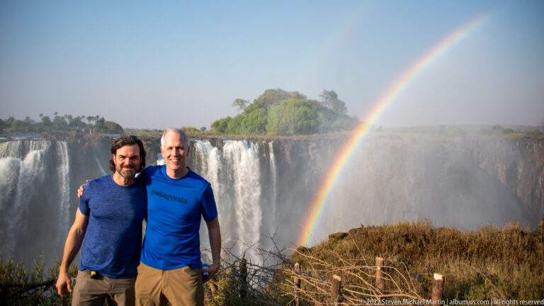 Two men standing together before Victoria Falls, with a rainbow, mist, green island, and overlook barrier in the background.