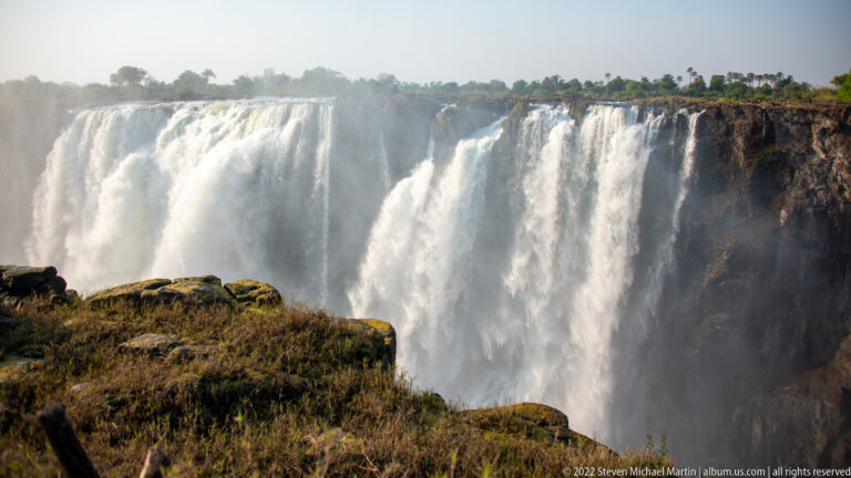 Massive white curtains of Victoria Falls dropping into dense spray, with low sunlit rock and dry vegetation in front.