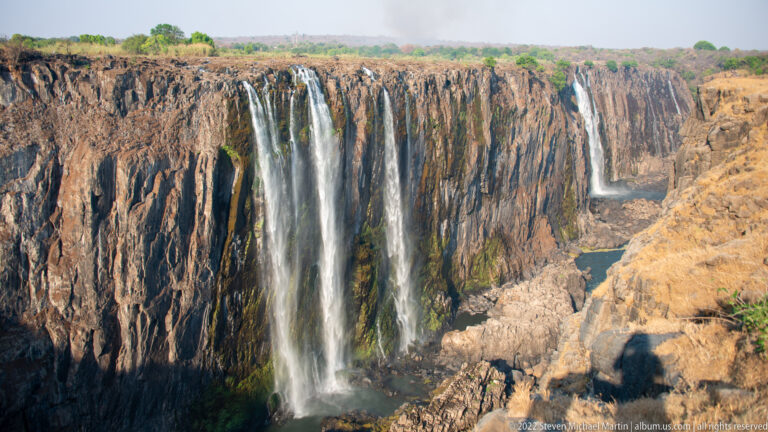 Several narrow waterfalls descending a largely exposed dark basalt cliff, with the river channel visible at the bottom of the gorge.