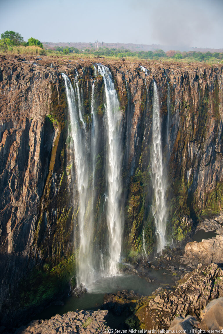Three main waterfall streams descending an exposed basalt cliff into a dark basin, with green staining along wetter rock channels.