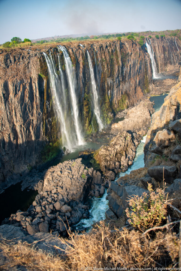 Wide rocky gorge below Victoria Falls with separated cascades on the far basalt wall and a narrow blue river channel below.