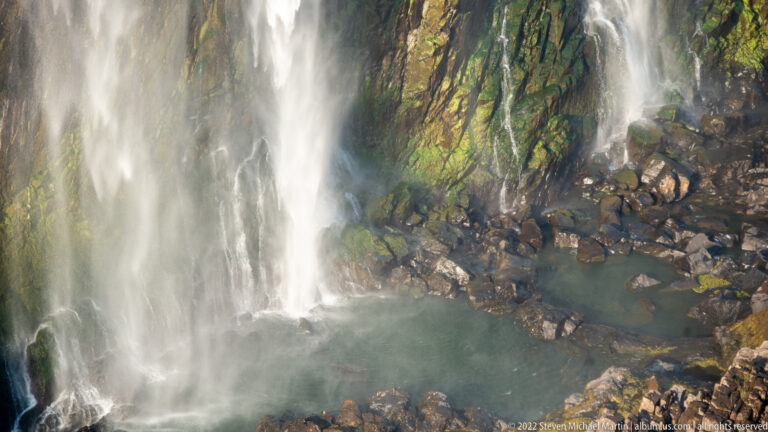 Close view of waterfall spray striking dark basalt boulders, with green wet rock and a small pool at the base.