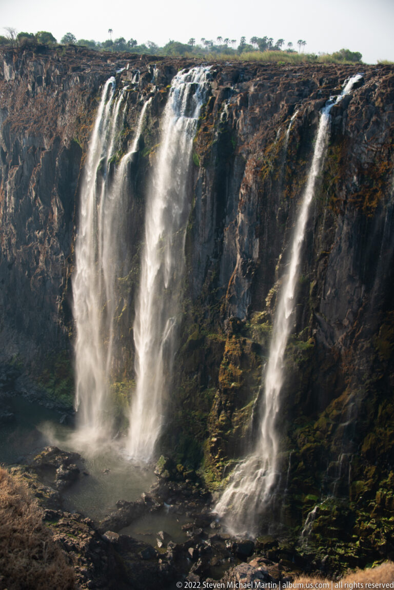 Thin white waterfall veils descending a broad dark basalt cliff, with wet ledges and shallow pools visible at the base.