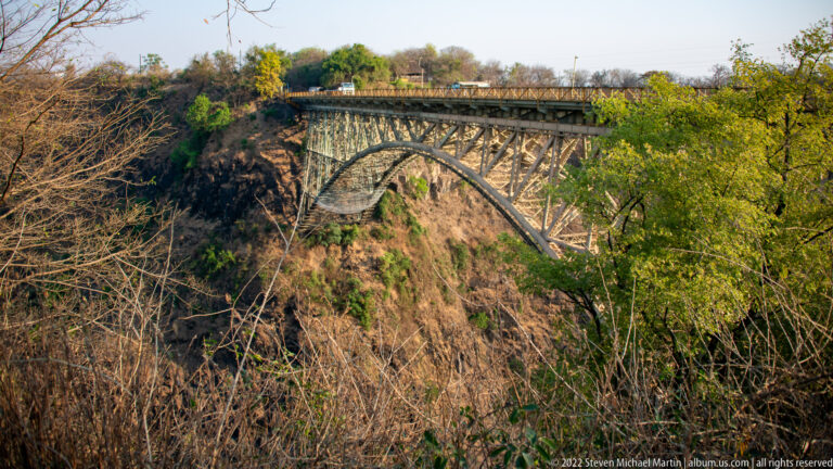 Steel arch Victoria Falls Bridge spanning the gorge, partly framed by dry scrub and green trees along the rocky rim.