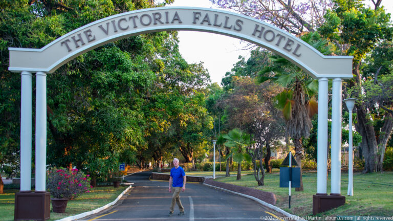 White Victoria Falls Hotel entrance arch over a tree-lined driveway with a pedestrian beneath it.
