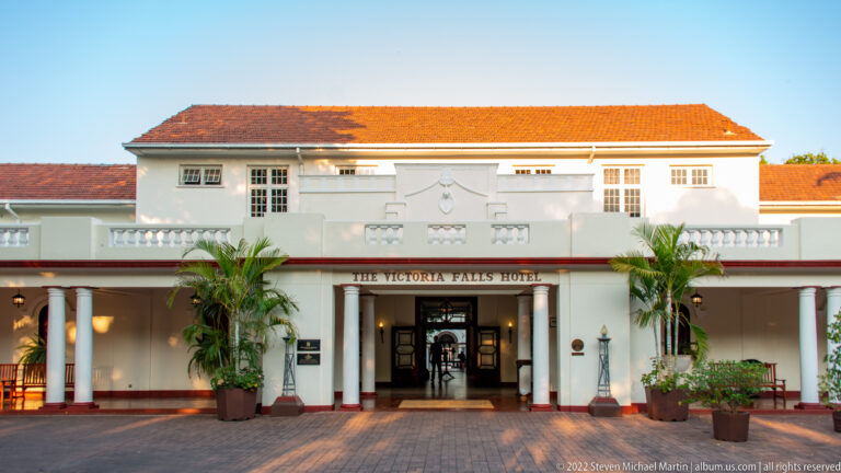 Symmetrical white façade of The Victoria Falls Hotel with red-tiled roof, columns, palms, and central entrance.