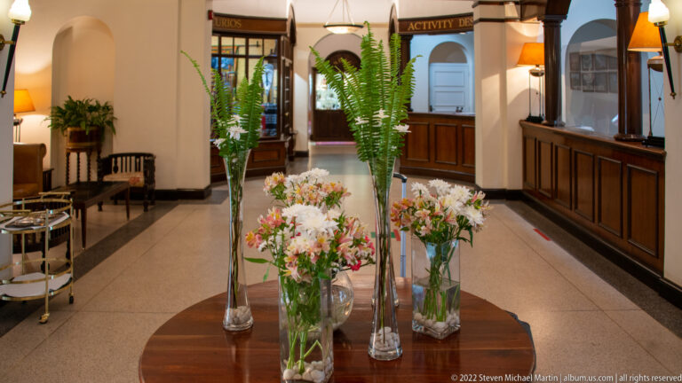 Hotel lobby corridor with tall flower arrangements, dark timber counters, polished floor, and activity-desk signage.