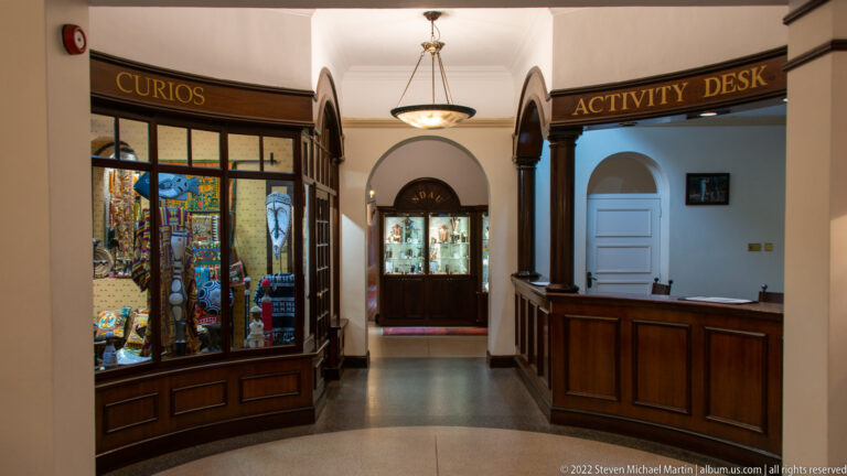 Narrow hotel corridor with dark wood display cases, arched openings, hanging light, and activity-desk counter.