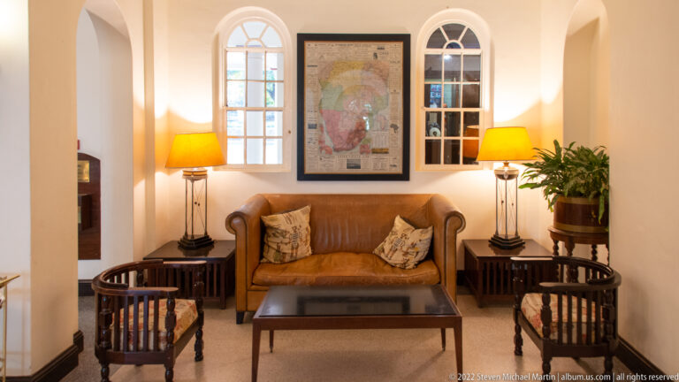 Hotel sitting alcove with tan leather sofa, wooden chairs, lamps, arched windows, and a framed map.