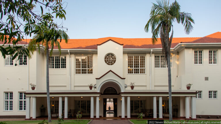 Garden-side white hotel façade with red roof, palms, gridded windows, circular window, and central arched entrance.