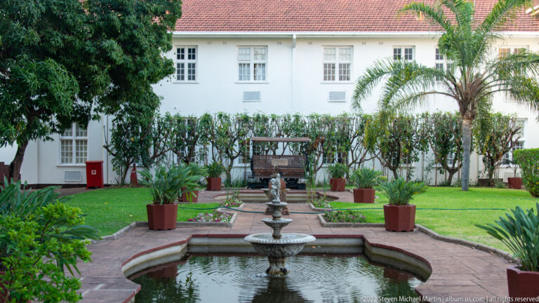 Formal hotel courtyard with tiered fountain, brick paths, potted plants, palms, lawn, and white building.