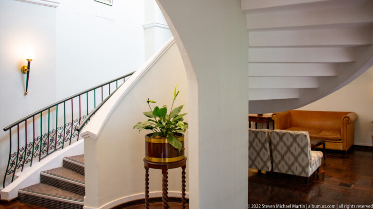 Curving white hotel staircase with black iron railing, potted plant, dark floor, and tan lounge seating.