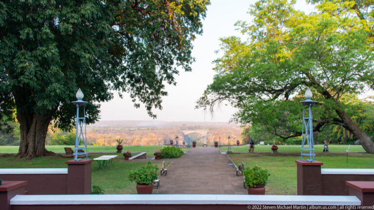 Straight garden path from the hotel terrace through lawns and mature trees toward a hazy distant landscape.