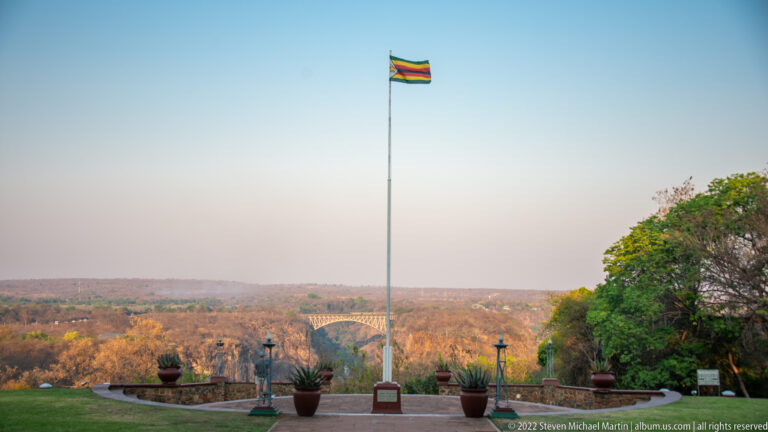 Zimbabwe flag flying on a tall pole above landscaped hotel grounds and a distant wooded horizon.