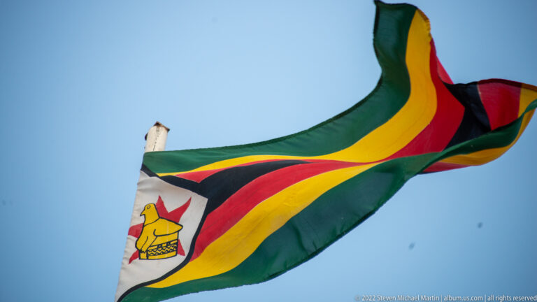 Zimbabwe national flag billowing against blue sky, with multicolored stripes and the white triangle partly visible.
