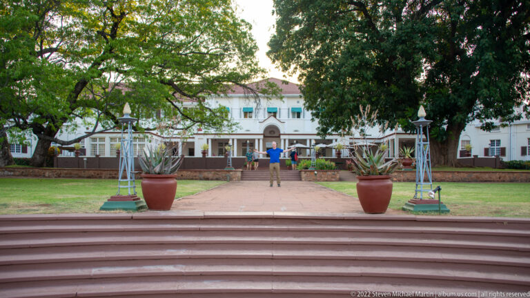 Broad steps and a straight garden path lead toward the white Victoria Falls Hotel between large potted plants and trees.