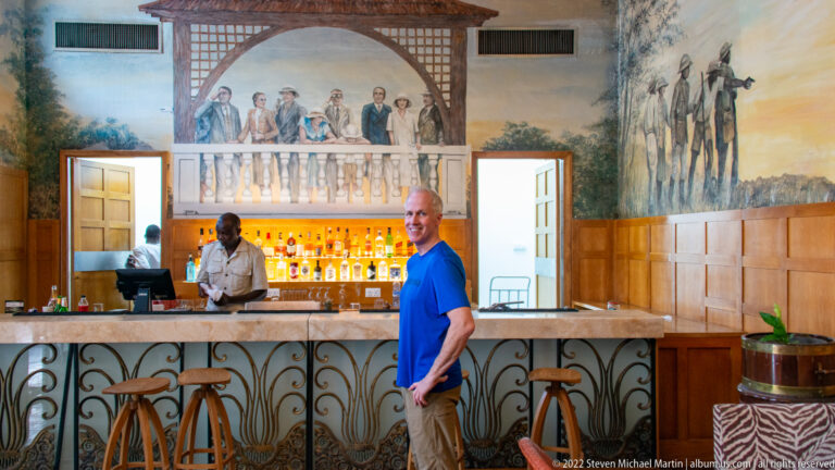 Hotel bar with illuminated bottles, ironwork counter, wall murals of period figures, a bartender, and a guest.
