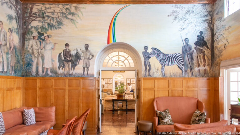 Hotel lounge with safari-themed murals, zebra figure, wood paneling, leather seating, and a centered arched doorway.