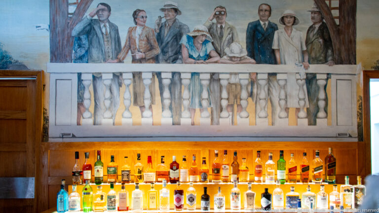 Illuminated rows of liquor bottles below a mural of period-dressed travelers standing behind a painted balustrade.