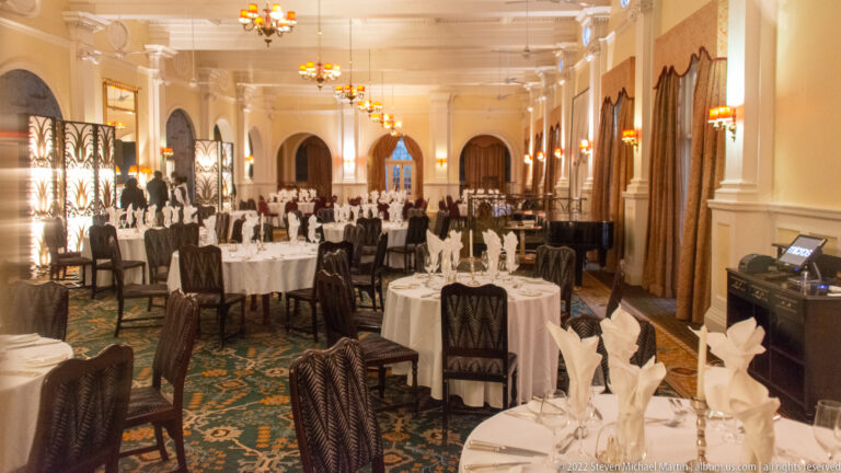 Long formal hotel dining room with white-clothed round tables, dark chairs, chandeliers, curtains, and arched openings.