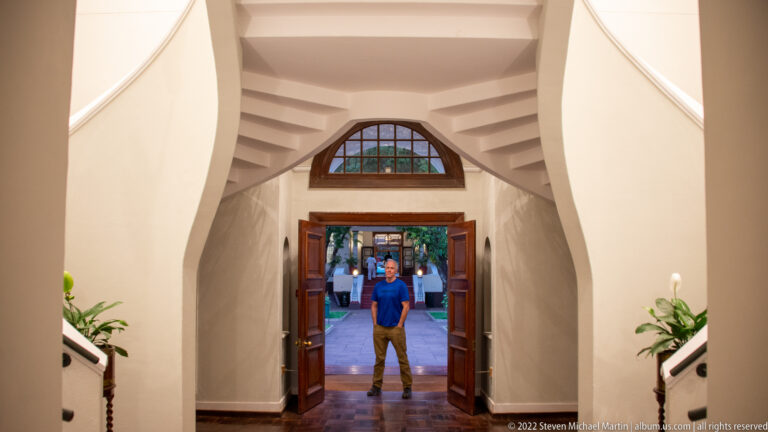 Person standing in an open doorway beneath a stained-glass transom, framed by white walls and symmetrical stairs.
