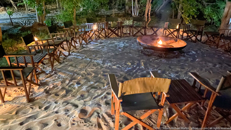 Burning circular fire bowl surrounded by safari chairs on pale sand beneath trees at night.