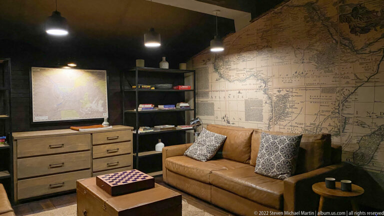 Tan leather sofa, trunk coffee table, shelving, and large map-covered wall in a compact safari lodge lounge.