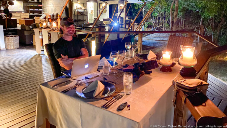 Traveler seated at a lantern-lit dinner table on a wooden deck with wine glasses, place settings, and a laptop.