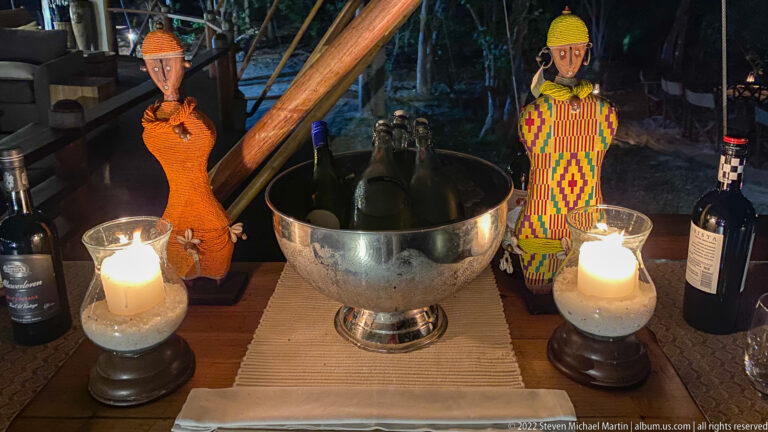 Silver bottle cooler between two decorative African figures and two glowing glass lanterns on a lodge table.