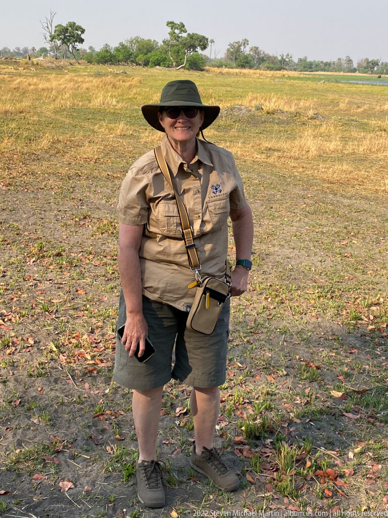 Safari traveler in khaki field clothing and a wide-brimmed hat standing on open floodplain ground with gear slung across the body.