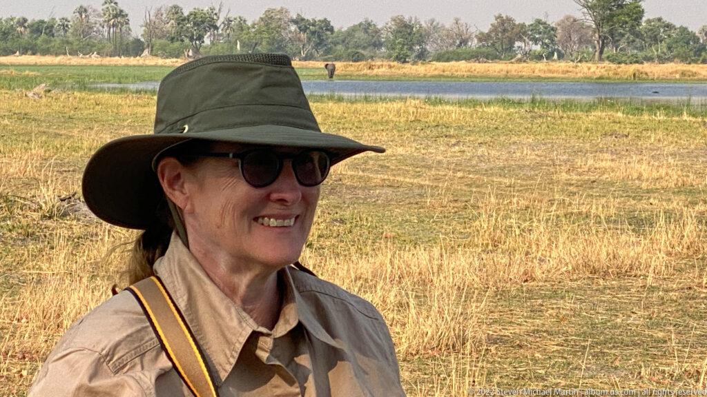 Safari traveler in a broad-brimmed hat and sunglasses standing before dry grass, a water channel, and distant woodland.