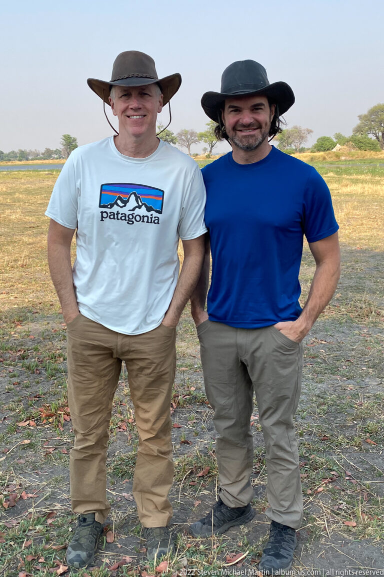 Two safari travelers in wide-brimmed hats standing together on open ground with a grassy floodplain behind.