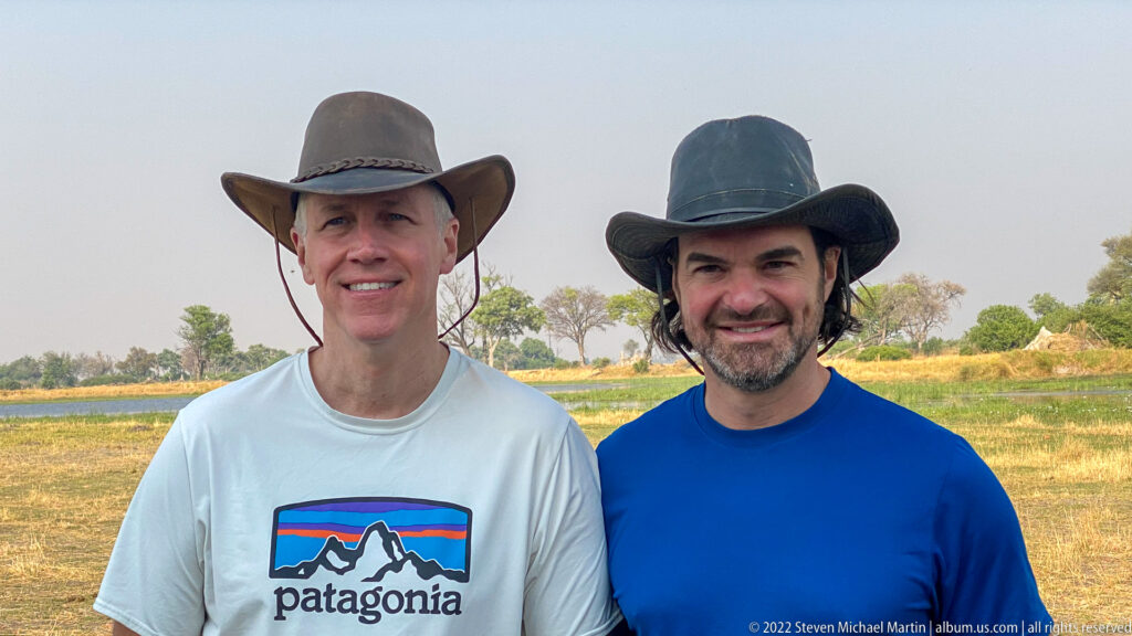 Close portrait of two safari travelers in hats with a wetland and scattered trees softly visible behind them.