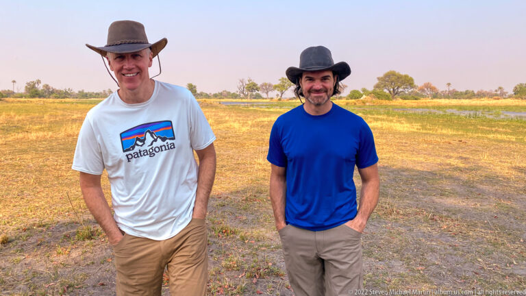 Two safari travelers standing full length on dry ground with warm-lit floodplain and trees in the background.