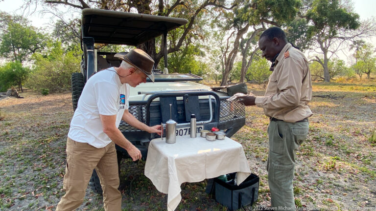 Safari guide and traveler preparing drinks at a small covered table beside an open game-drive vehicle under trees.