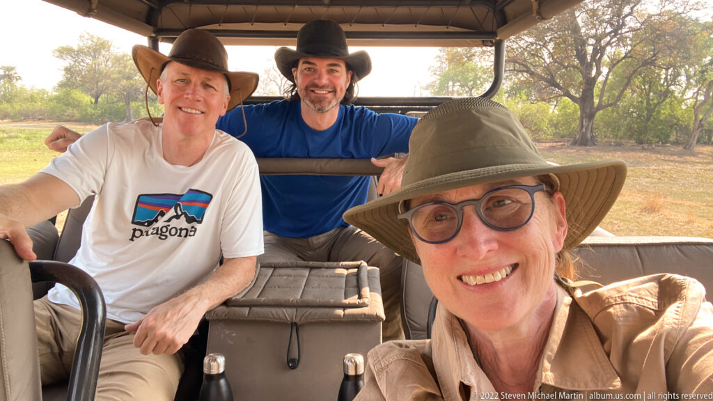 Three safari travelers seated together inside an open game-drive vehicle with woodland visible through the sides.