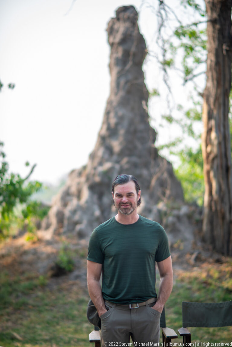 Safari traveler standing before a very large irregular termite mound between mature trees.